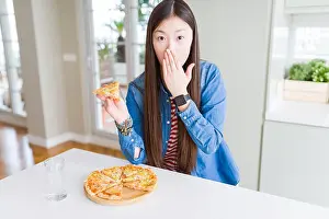 Shocked Prints: Beautiful Asian woman eating a slice of cheese pizza cover mouth with hand shocked with shame for mistake