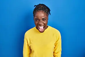 Beautiful black woman standing over blue background angry and mad screaming frustrated and furious