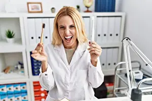 Dentist Prints: Beautiful blonde dentist woman holding electric toothbrush and normal toothbrush angry and mad screaming frustrated