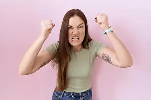 Beautiful brunette woman standing over pink background angry and mad raising fists frustrated