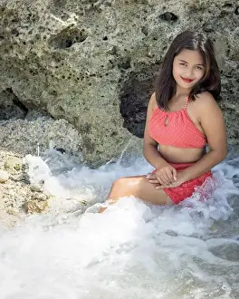 A beautiful Filipina in a red swimsuit smiling and sitting in beach water with a rock in the back