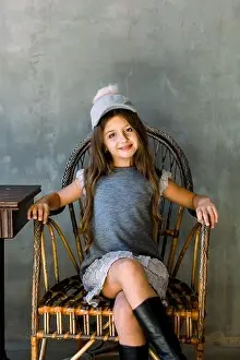 Beautiful little girl with brown long hair and blue eyes sitting in an armchair dressed in gray dress and a hat
