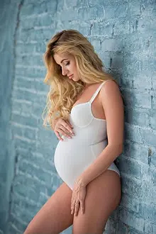 Abdomen Prints: Beautiful pregnant blonde woman in a white swimsuit stands near a gray brick wall and looks at her belly