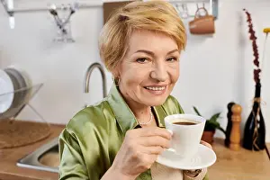 Contentment Prints: A beautiful senior woman holds a cup of coffee while smiling warmly in her bright kitchen