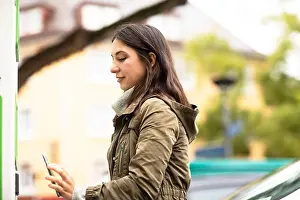 Beautiful Woman Prints: Beautiful woman using smart phone for renting electric car at charging station