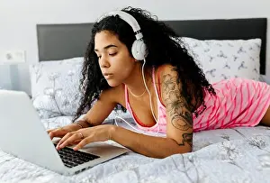 Millennials Prints: Beautiful young woman lying in bed at home wearing headphones and using laptop