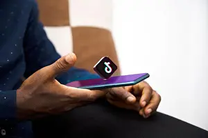 Boredom Prints: Black Hand holding mobile phone with Tiktok logo hovering on screen in living room
