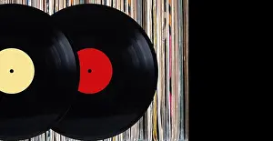 Analogue Prints: Two black vinyl records with color labels in front of shelf of many close standing vinyl discs in old color covers over