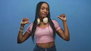 Braids Prints: Black woman in pink crop top with headphones around neck points thumbs to chest against a studio blue backdrop