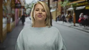 Inquiry Prints: Blonde woman wearing light sweater pointing finger at blurred figures on bustling city street; curiosity