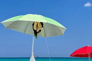 Summer Vacation Prints: Brightly colored beach umbrella on a sandy beach overlooking calm turquoise water and a yacht in the distance