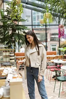 Temptation Prints: brunette young woman with long hair in beige leather jacket and denim jeans standing near cake display with pastry