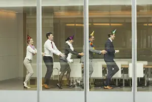 Coworker Prints: Business people wearing party hats dancing in conga line at conference room window