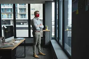 Crossed Prints: Caucasian man wearing face mask with arms crossed looking out of window at modern office