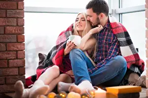 Plaid Prints: Charming young couple man and woman bask in a blanket while sitting on a windowsill with a winter look