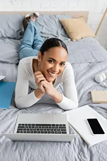 Blouse Prints: Cheerful african american student lying near gadgets and notebooks on bed, stock image