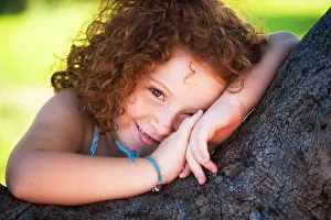 Redhead Prints: Cheerful girl with curly red hair smiling while leaning on a tree outdoors