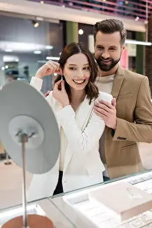 Customers Prints: Cheerful man hugging girlfriend wearing earring near mirror in jewelry shop