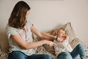 Cheerful teen girl sitting on bed and tickling laughing sister while playing together at home during weekend