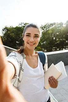 Selfie Prints: Cheerful young student girl carrying backpack outdoors, holding books, taking selfie