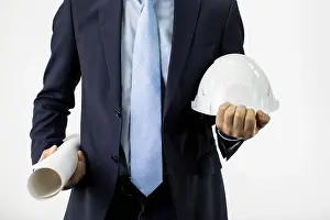 Discussing Prints: Close up engineer in formal suit holds hard safety helmet hat and blueprint roll