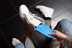 Barefoot Prints: Close up of female hands putting custom orthopedic insoles inside white sneakers over black floor tiles