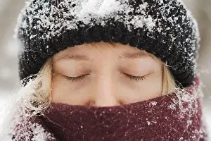 Beautiful Woman Prints: Close-up portait of a blonde woman in beanie hat and nose covered in scarf stands with eyes closed in winter snowfall