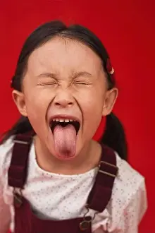 Eyes Closed Prints: Close-up portrait of funny little Asian girl with two ponytails grimacing on camera sticking with her tongue out