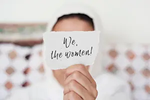 Veil Prints: closeup of a young woman indoors wearing a white hijab showing a piece of paper in front of her face with the text we