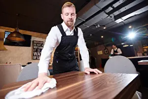 Waiter Prints: Content handsome young waiter with beard wearing white shirt and apron standing at wooden table