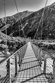 Boulders Prints: Contrast BW hanging foot walking suspension bridge across Hooker river in scenic mountain valley of New Zealand on a