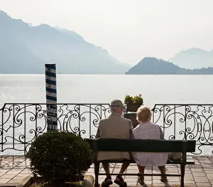 Lake Como Prints: Couple sitting on the lake front promenade enjoying the sunshine and views of lake Como
