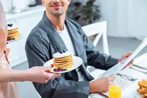 Partial Prints: cropped view of woman holding plates with pancakes while man reading newspaper during breakfast in kitchen