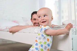 Headboard Prints: Cute little baby is standing on the edge of the bed holding on to the headboard against the background of his parents