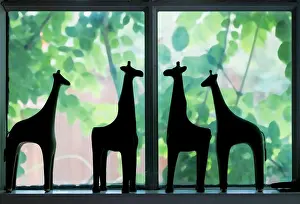 Calming Prints: Cute silhouettes of giraffe figures standing on a window ledge with blurred greenery and flowers showing through