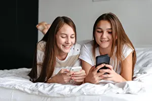 Texting Prints: Two cute smiling teengae girls laying on bed, holding mobile phones at home