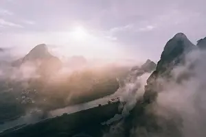 Humid Prints: Drone view of calm river and rocky dark mountains in morning mist with flowing clouds under bright sky and sun
