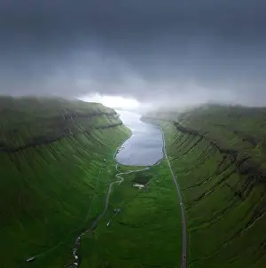 Drone view of curvy roads going on green mossy fjord near calm sea water against gray overcast sky on stormy day