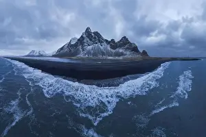 From above drone view of rocky mountains covered with snow located near waving seawater near black sand beach in
