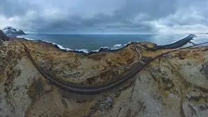 Drone view of sea waves washing rocky coast with asphalt road going through mountainous terrain during daytime in