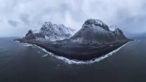 Drone view of sea waves washing stony beach with snowy mountains against cloudy sky in Iceland