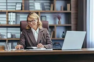 Business Attire Prints: Elegant senior woman in business attire smiles while working at her laptop in a well-appointed home office setup