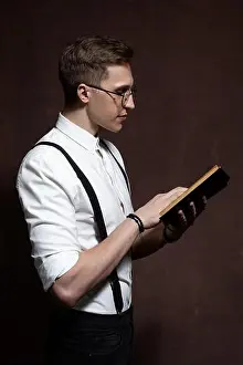 elegant young man in round glasses, suspenders, in a white shirt reads a book
