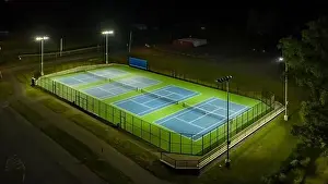 Evening aerial video of outdoor blue tennis courts with pickleball lines with lights turned on