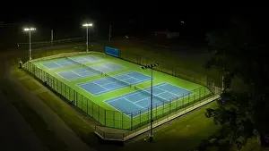 Evening aerial video of outdoor blue tennis courts with pickleball lines with lights turned on