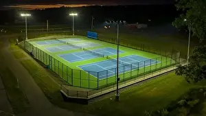 Evening aerial video of outdoor blue tennis courts with pickleball lines with lights turned on