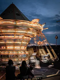 Spectators Prints: In the evening the carousel with many lights turns fast in front of steady spectators
