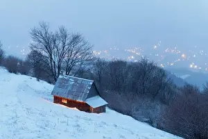 Fantastic winter landscape with glowing wooden house against the backdrop of glowing city lights in fog. Cozy cabin in Carpathian mountains
