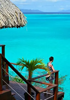 Honeymoon Prints: Female tourist on the balcony of an overwater bungalow with thatched roof in a luxury resort looking over the Bora Bora