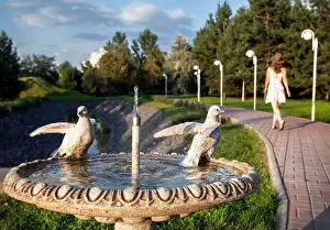 Honeymoon Prints: Fountain with dove statue and woman in white dress walking in the summer park in Almaty
